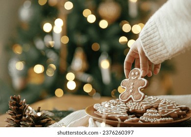 Merry Christmas! Hand holding gingerbread man cookie with icing on background of cookies in plate on table against christmas tree golden lights. Atmospheric Christmas holidays, family time