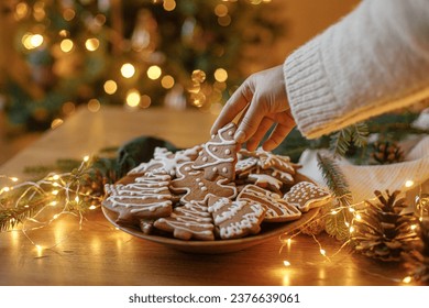 Merry Christmas! Hand holding gingerbread cookie with icing on background of cookies in plate on table against christmas tree golden lights. Atmospheric Christmas holidays, family time - Powered by Shutterstock