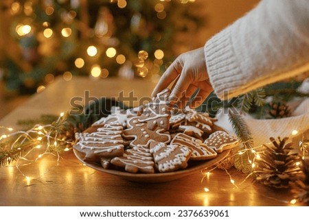 Merry Christmas! Hand holding gingerbread cookie with icing on background of cookies in plate on table against christmas tree golden lights. Atmospheric Christmas holidays, family time