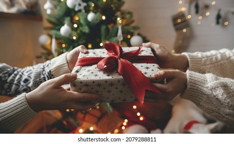 Merry Christmas! Hands exchanging christmas gift with red bow on background of christmas tree with lights. Stylish couple holding present with red ribbon close up in festive decorated room - Powered by Shutterstock