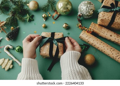 Merry Christmas! Hands wrapping stylish christmas gift, golden wrapping paper, green ribbon and festive decorations on green background. Wrapping christmas gifts concept. Happy Holidays