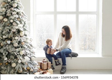 Merry Christmas And Happy Holidays! Cheerful Mom And Her Cute Baby Boy With Gift Box Near Window And Christmas Tree Indoors .