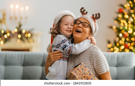 Merry Christmas and Happy Holidays! Cheerful grandma and her cute grand daughter girl exchanging gifts. Granny and little child having fun near tree indoors. Loving family with presents in room.
