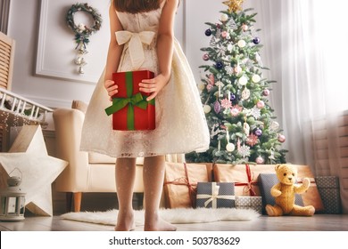 Merry Christmas And Happy Holidays! Cheerful Cute Little Child Girl With Present. Kid Holds A Gift Box Near Christmas Tree Indoors.