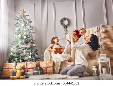 Merry Christmas and Happy Holidays! Cheerful mom and her cute daughters girls exchanging gifts. Parent and two little children having fun and playing together near Christmas tree indoors.