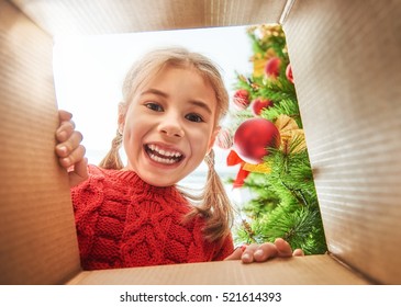 Merry Christmas And Happy Holidays! Cheerful Cute Child Girl Opening A Christmas Present. Little Kid Having Fun Near Christmas Tree Indoors. View From Inside Of The Box.