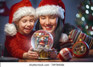 Merry Christmas and Happy Holidays! Cheerful mom and her cute daughter girl with snow globe. Parent and little child having fun near tree indoors. Loving family in room at home. - Powered by Shutterstock