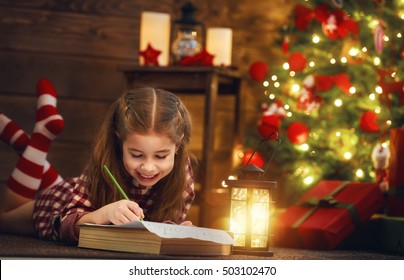 Merry Christmas And Happy Holidays! Cute Little Child Girl Writes The Letter To Santa Claus Near Christmas Tree Indoors. 