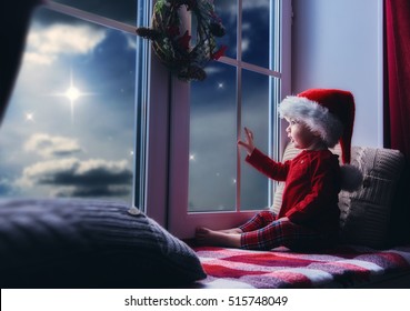 Merry Christmas And Happy Holidays! Cute Little Baby Girl Sitting By The Window And Looking At The Christmas Star. Room Decorated On Christmas. Kid Enjoys The Night Sky.