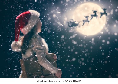 Merry Christmas and happy holidays! Cute little child girl with xmas present. Santa Claus flying in his sleigh against moon sky. Kid enjoy the holiday. Portrait kid with gift on dark background. - Shutterstock ID 519218686