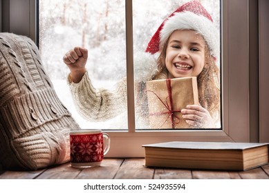 Merry Christmas And Happy Holidays! Cute Little Girl Looking In Window, Standing Outdoors On Winter Forest Background. Child Holding Present Gift Box.