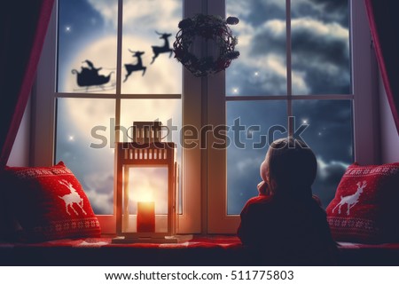 Merry Christmas and happy holidays! Cute little child girl sitting by window and looking at Santa Claus flying in his sleigh against moon sky. Room decorated on Christmas. Kid enjoy the holiday. 