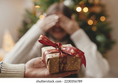 Merry Christmas and Happy Holidays! Hand holding christmas gift box on background of happy woman closing eyes waiting for surprise. Exchanging presents at christmas tree in atmospheric room