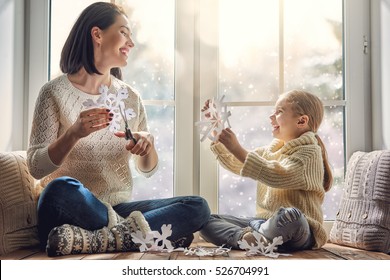 Merry Christmas and happy holidays! Happy loving family sitting by the window and making paper snowflakes for decoration windows. Mother and child creating decorations.