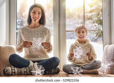 Merry Christmas And Happy Holidays! Happy Loving Family Sitting By The Window And Making Paper Snowflakes For Decoration Windows. Mother And Child Creating Decorations.