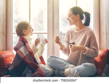 Merry Christmas And Happy Holidays! Happy Loving Family Sitting By The Window And Making Paper Snowflakes For Decoration Windows. Mother And Child Creating Decorations.