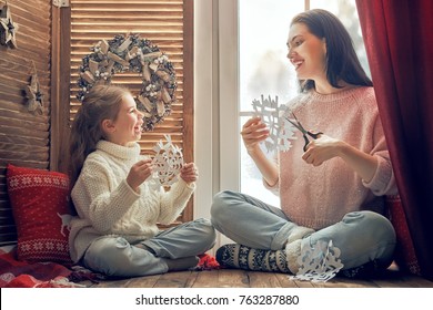 Merry Christmas And Happy Holidays! Happy Loving Family Sitting By The Window And Making Paper Snowflakes For Decoration Windows. Mother And Child Creating Decorations.