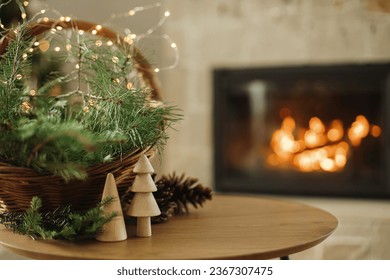 Merry Christmas and happy holidays! Stylish wicker basket with fir branches, wooden trees, pine cones on table against burning fireplace. Modern rustic eco friendly decor in living room - Powered by Shutterstock