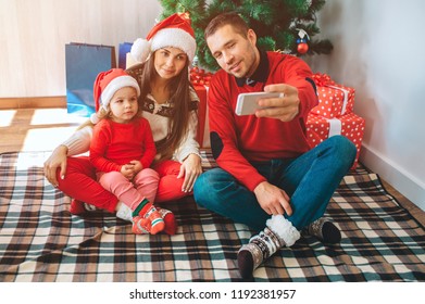 Merry Christmas And Happy New Year. Young Man Sits Besides Woman And Child. He Holds Phone And Takes Selfie. Woman And Kid Lok At It And Pose. Family Wears Christmas Clothes And Hats.