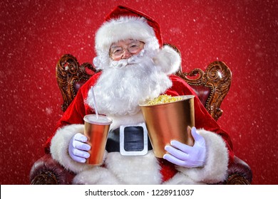 Merry Christmas and Happy New Year. Santa Claus sitting on his armchair and eating popcorn and drinking soda. Entertainment and cinema concept. Red background.