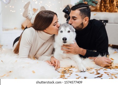 Merry Christmas And Happy New Year. Young Couple And A Cute Dog. Couple Spending Christmas Eve At Home. Couple Kisses Dog. Christmas Holidays Concept.