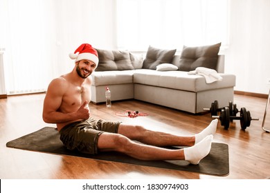 Merry Christmas And A Happy New Year Guy In Santa's Christmas Red Hat Has A Workout At Home. He Sits On A Topless Rug And Shows A Thumbs Up.