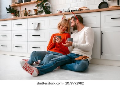 Merry Christmas and a Happy New Year! Young loving couple with reindeer antlers in the kitchen drink coffee with cookies sitting on the floor in anticipation of the holiday. - Powered by Shutterstock