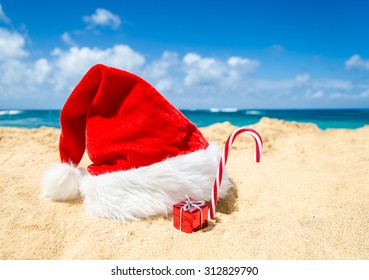 Merry Christmas And Happy New Year Background With Santa Claus Hat, Candy And Gift On The Tropical Beach Near Ocean In Hawaii