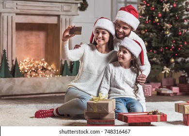 Merry Christmas And Happy New Year! Beautiful Mom, Dad And Daughter In Santa Hats Are Doing Selfie Using A Smart Phone And Smiling While Celebrating