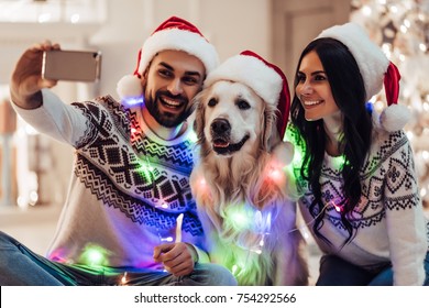 Merry Christmas and Happy New Year! Happy couple with dog labrador retriever waiting for the New Year in Santa Claus hats while sitting near beautiful Christmas tree at home. Smiling and making selfie - Powered by Shutterstock