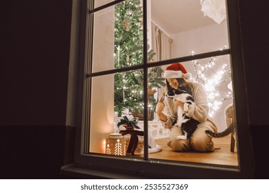 Merry Christmas! Happy woman in santa hat hugging cute cat in window on background of stylish gifts and christmas tree in festive living room with lights. Atmospheric christmas eve at home - Powered by Shutterstock