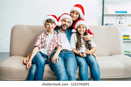 Merry Christmas! Happy Young Family In Red Santa Hats Are Hugging Each Other And Spending Time In Living Room While Sitting On The Couch