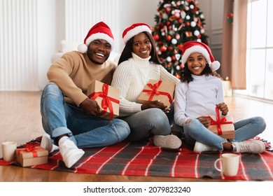 Merry Christmas. Portrait Of Happy Loving Black Family In Red Santa Claus Hat Celebrating Winter Holidays Together, Holding Boxes Sitting On Floor Blanket In Living Room, Posing Looking At Camera