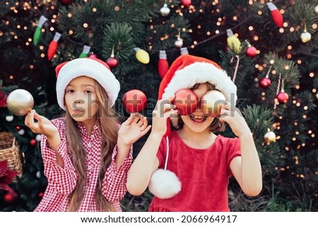 Merry Christmas. Portrait of two happy funny children girls in Santa hat with Christmastree toys near face. No face. Happy Holidays. Fairy Magic. Happy kids enjoying holiday.