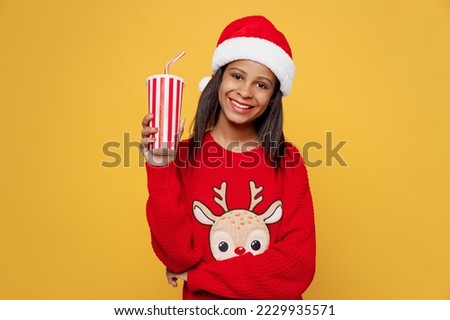 Merry fun little kid teen girl 13-14 years old wear red xmas sweater with deer Santa hat posing hold cup of soda pop cola water isolated on plain yellow background. Happy New Year 2023 holiday concept