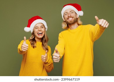 Merry fun young couple two friends man woman wear sweater Santa hat posing showing thumb up like gesture wink isolated on plain green background. Happy New Year celebration Christmas holiday concept - Powered by Shutterstock