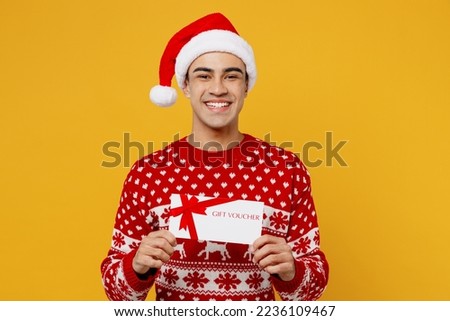 Merry fun young man wear red knitted christmas sweater Santa hat posing hold gift certificate coupon voucher card for store isolated on plain yellow background. Happy New Year 2023 celebration concept