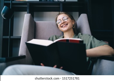 Merry And Glad Woman In Glasses Sitting In Comfortable Armchair In Library And Laughing While Reading Funny Story In Book