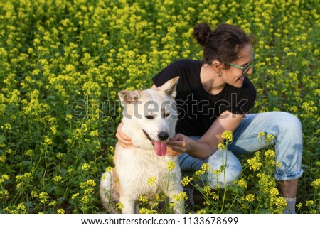 A merry laughing happy curly-haired brunette girl with glasses hugs a red-haired smiling akita inu dog in a field among yellow wild flowers on a sunny summer day.