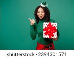 Merry little kid teen girl wears hat casual clothes posing hold present box with gift ribbon bow point aside isolated on plain green background. Happy New Year celebration Christmas holiday concept