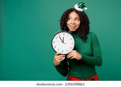 Merry little kid teen girl wear hat casual clothes posing hold in hand clock look aside on area isolated on plain green background studio portrait. Happy New Year celebration Christmas holiday concept - Shutterstock ID 2396398861