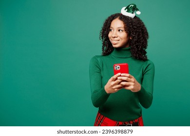 Merry little kid teen girl wear turtleneck hat casual clothes posing hold in hand use mobile cell phone look aside isolated on plain green background studio portrait. Happy New Year holiday concept - Shutterstock ID 2396398879