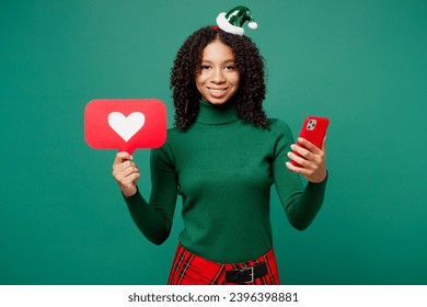Merry little kid teen girl wear casual clothes posing use mobile cell phone heart form like icon sign from social network isolated on plain green background. Happy New Year Christmas holiday concept - Shutterstock ID 2396398881