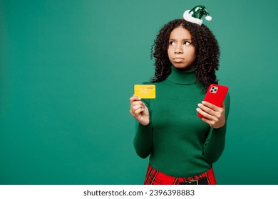 Merry little kid teen girl wears hat casual clothes posing using mobile cell phone credit bank card shopping online isolated on plain green background studio. Happy New Year Christmas holiday concept - Shutterstock ID 2396398883
