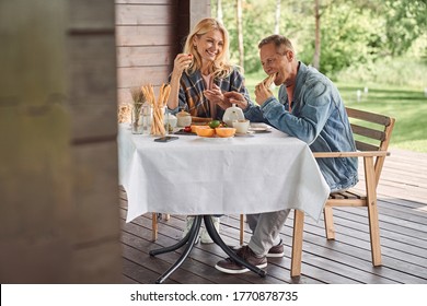 Merry man and woman are enjoying hot tea and sandwiches while sitting at table on terrace