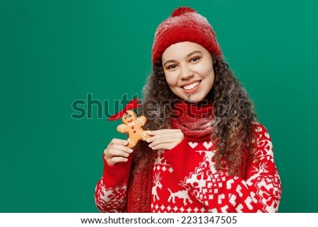 Merry young fun woman wearing red warm cozy knitted sweater hat posing hold ginger cookie man isolated on plain dark green background studio portrait. Happy New Year 2023 celebration holiday concept