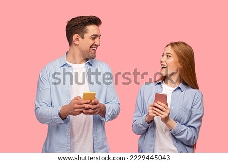 Merry young girlfriend and boyfriend in similar clothes using cellphones and looking at each other with smile against pink background