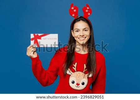 Merry young Latin woman wear red Christmas sweater decorative fun deer horns on head posing hold gift coupon voucher card for store isolated on plain blue background. Happy New Year holiday concept