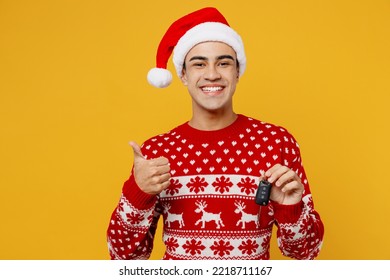 Merry young man wear red knitted christmas sweater Santa hat posing hold car key fob keyless system show thumb up isolated on plain yellow background. Happy New Year 2023 celebration holiday concept