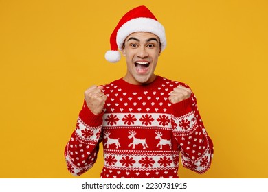 Merry young man wear red knitted christmas sweater Santa hat posing doing winner gesture celebrate clenching fists say yes isolated on plain yellow background. Happy New Year 2023 celebration concept - Powered by Shutterstock